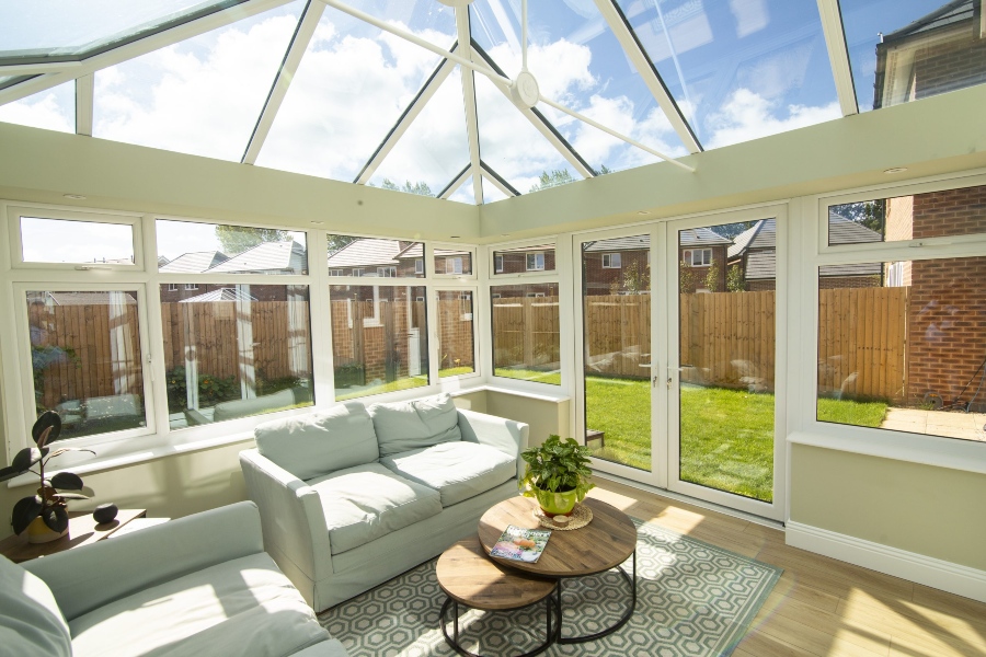 A light-filled Hup! sunroom with a pitched glass roof, white framing, and a ceiling fan. The room features two light green sofas facing each other with two round wooden coffee tables in the middle. Large windows and a set of glass doors look out onto a back garden and neighboring houses.
