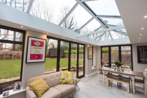 A large, open-plan living and dining space with a modern, black-framed Hup! glass roof and two sets of large black bi-fold doors that open onto a garden. The room has a beige sofa with yellow throw pillows and a dining table with a bench and chairs. The floor is light wood, and the walls are painted a neutral grey.