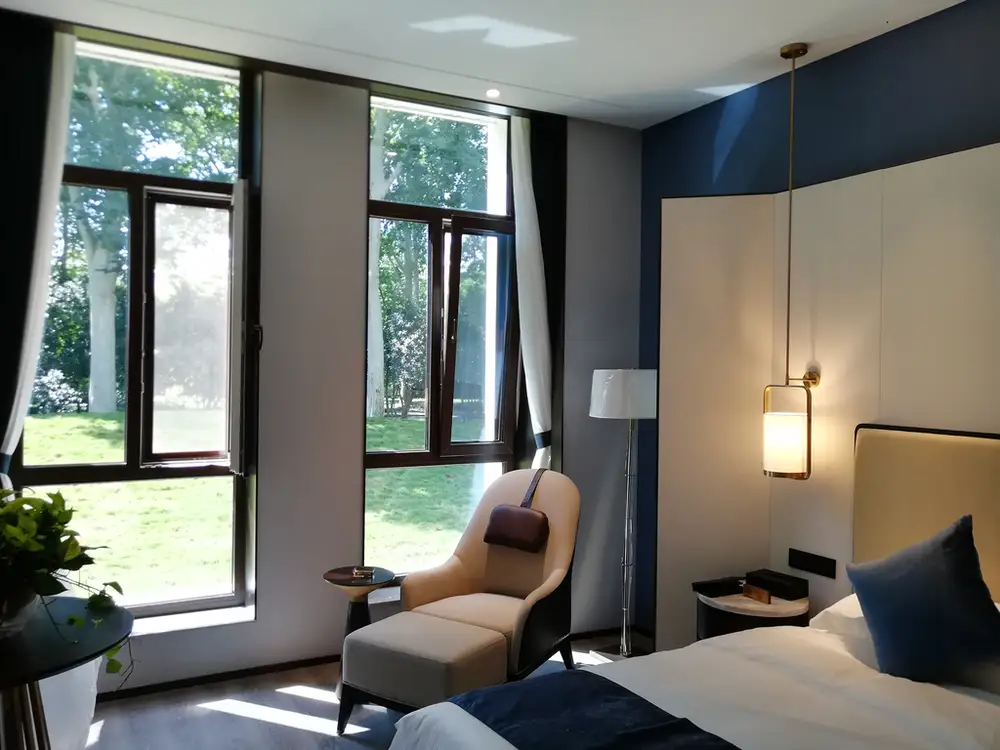 A well-lit, modern bedroom with dark blue and white walls. Two large, dark-framed tilt and turn windows on the left look out onto a lush green lawn and trees. A beige chaise lounge with a handbag on it sits between the windows.