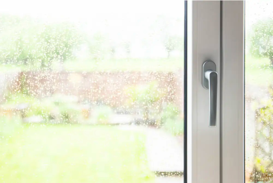 A close-up, eye-level shot of a modern tilt & turn window on a rainy day. The window is made of white UPVC with a silver handle on the right side. The glass is covered in small raindrops, causing the green and brown garden outside to appear blurry and out of focus.