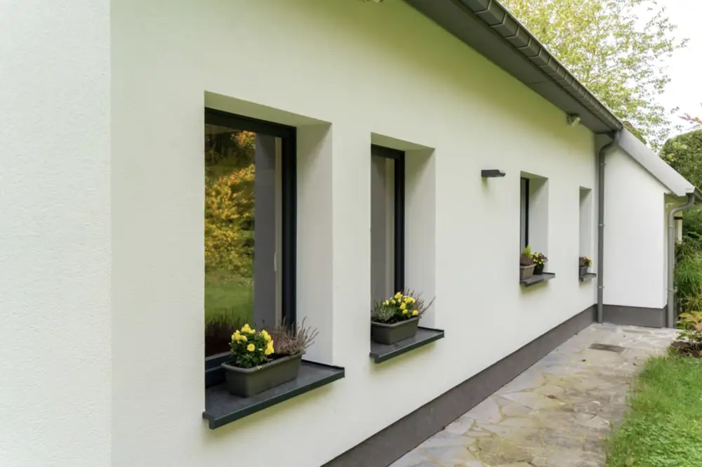 A modern, single-story house with a white rendered facade and dark grey trim. The side of the house features four rectangular tilt and turn windows with black frames and dark grey sills, each holding a window box with yellow flowers.